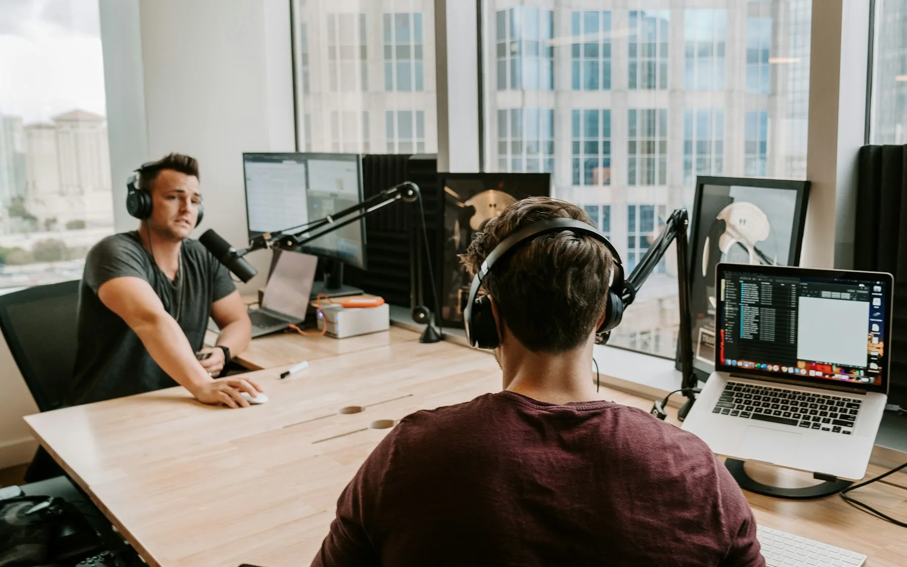 Doi oameni la o masă de podcast, două microfoane pe brațe articulate, laptop deschis